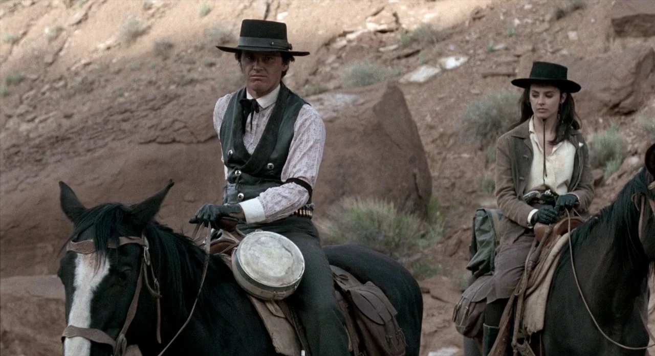 The Shooting (1966) movie still: medium — Two individuals on horseback traverse a dry, rocky desert terrain with reddish stone formations. They…