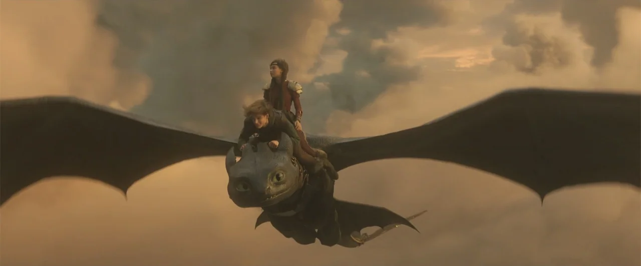 How to Train Your Dragon (2025) movie still: wide — A dark dragon flies through a vast expanse of golden, sunset-lit clouds. Two young riders are…