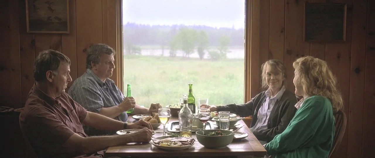 In the Bedroom (2001) movie still: medium — A wooden cabin dining room with wood-paneled walls overlooks a foggy green field. Four people sit around…