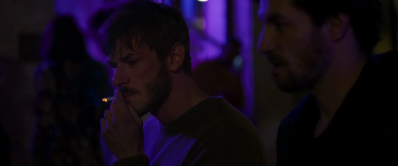 More Than Ever (2022) movie still: medium — A dimly lit nightlife setting with deep violet neon illumination throughout the space. Two men stand in…