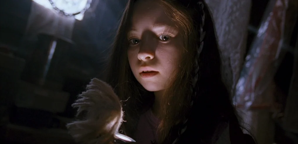 Tideland (2005) movie still: closeup — A dark, cluttered interior space with plastic-covered objects in the background. A young girl shines a small…