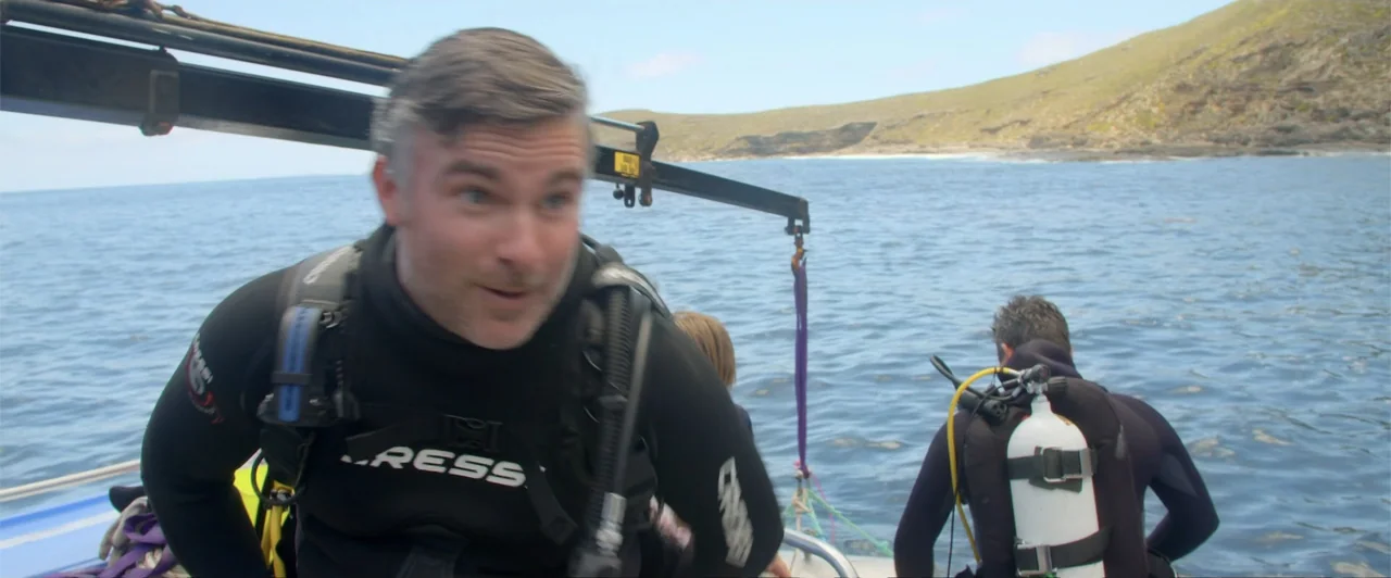 Lair of the Great White (2024) movie still: medium — A view from a boat deck on open water near a rocky coastline. A man in a diving suit looks…