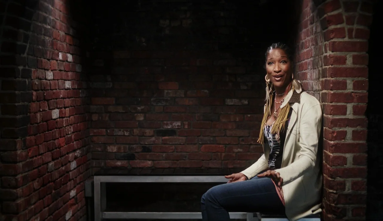 13th (2016) movie still: medium — A woman sits inside an enclosed area built of weathered dark red brick masonry. The woman is seated on a bench…