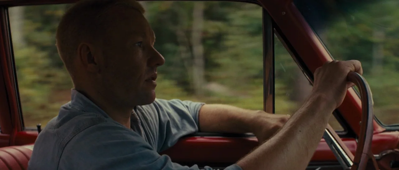 Loving (2016) movie still: medium — A man drives a red vehicle through a forested landscape during daylight hours. The driver holds the steering…