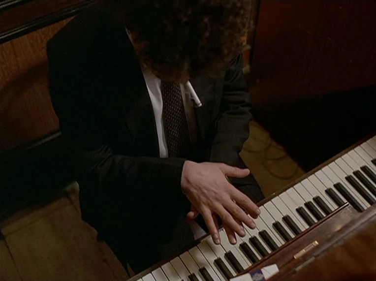 Detective (1985) movie still: medium — A dark, dimly lit room features a piano and wooden paneled walls. A man in a suit plays the instrument while…