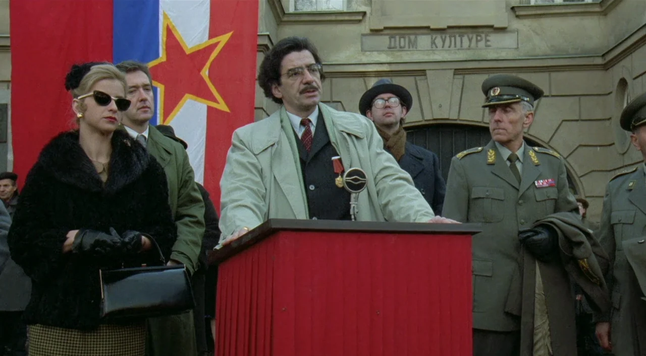 Underground (1995) movie still: medium — A man speaks at a podium in front of a building labeled Dom Kulture. A red flag with a yellow star is…