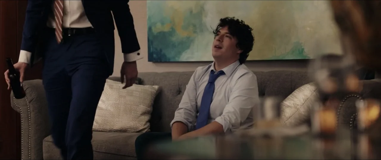 Plus One (2019) movie still: medium — A modern living room features a grey sofa placed in front of an abstract painting. A man sits on the sofa…