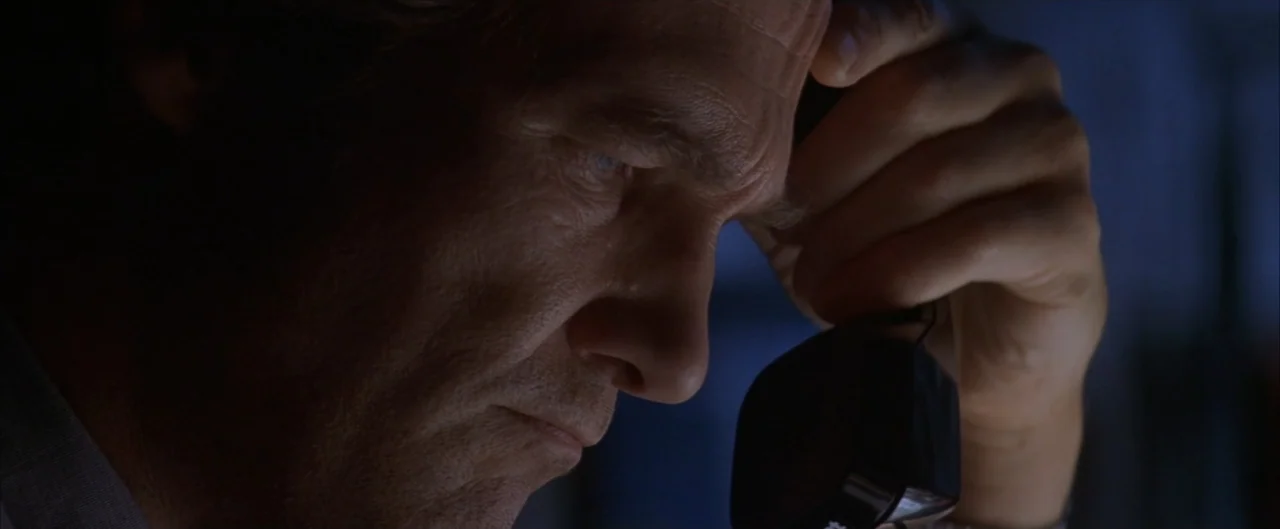 Arlington Road (1999) movie still: closeup — A dark indoor setting with limited illumination on the man's face. A man in his fifties listens to a…