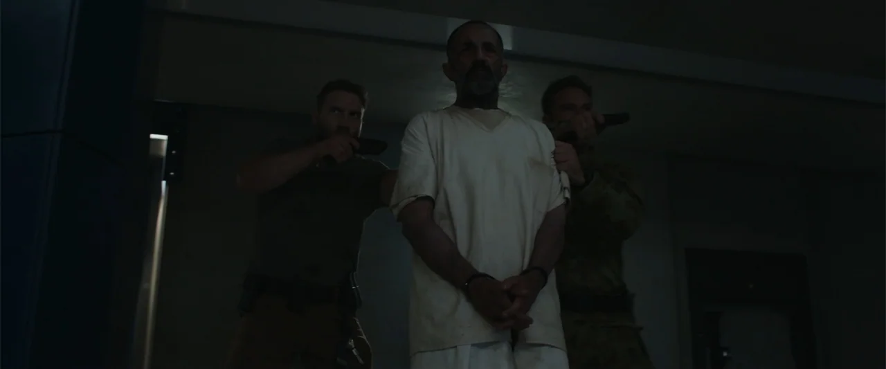 Black Site (2022) movie still: medium — A dimly lit room with plain walls and industrial ceiling details. Two armed men point handguns at a…