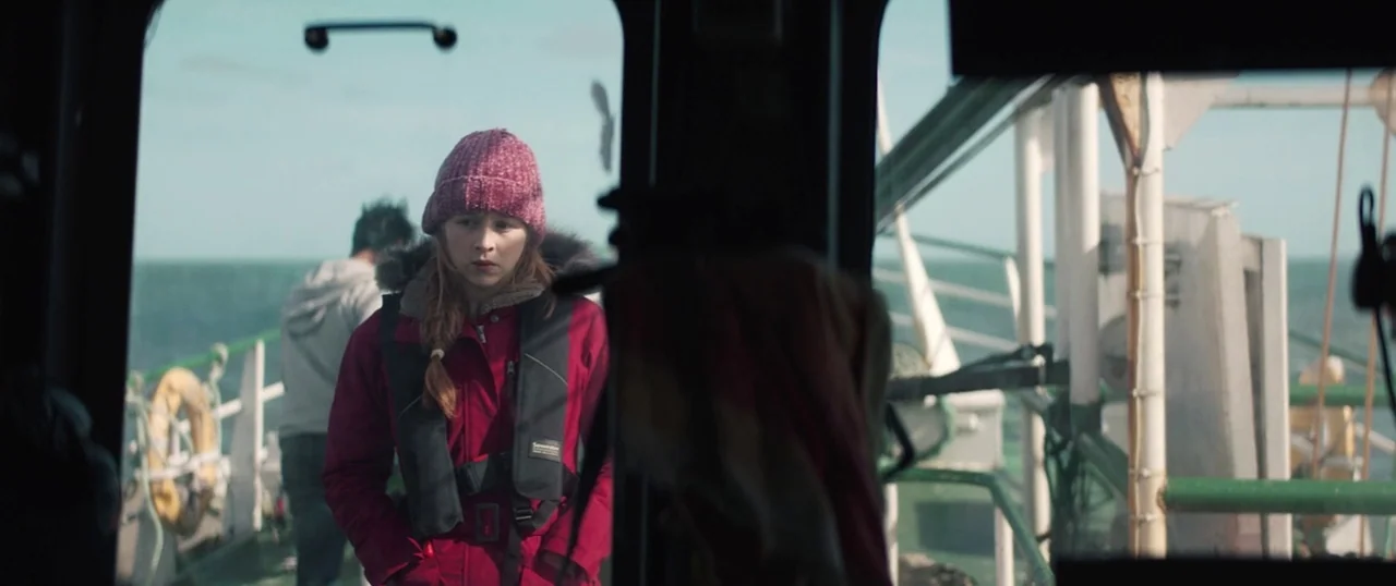 Sea Fever (2019) movie still: medium — The deck of a large vessel displays metal railings and a lifebuoy ring. A woman in a red jacket and beanie…
