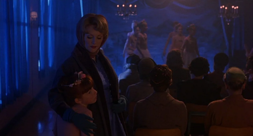 Far From Heaven (2002) movie still: medium — A dimly lit auditorium features a stage with young dancers under soft light. A woman in a dark coat and…