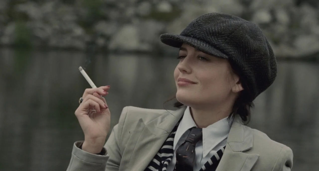 Cracks (2009) movie still: medium — A woman stands near a calm lake shore with a rocky, blurred background. She wears a formal blazer and cap while…