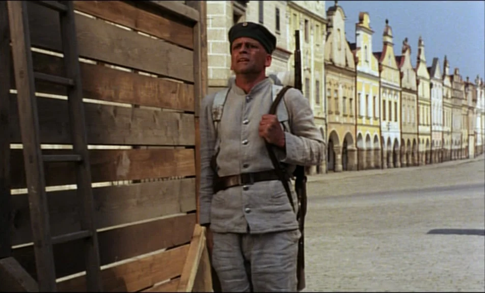 Woyzeck (1979) movie still: medium — A European town square with a row of pastel-colored historic buildings with arched ground-level walkways. A…