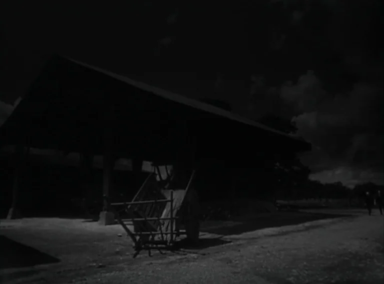 Head Against The Wall (1959) movie still: wide — An open wooden barn structure with a dirt floor stands under a cloudy sky. A simple wooden cart…