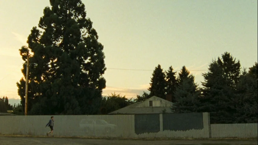 Wendy and Lucy (2008) movie still: wide — A suburban street scene with a long metal fence and several large pine trees. A lone boy walks along the…
