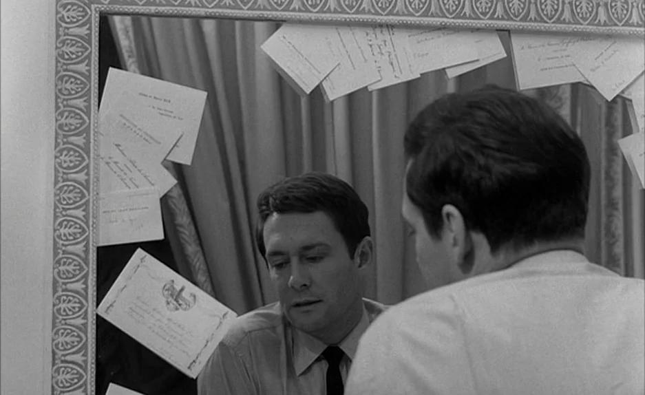 Le Feu Follet (1963) movie still: medium — A man's reflection is visible in a mirror framed by several white paper documents. The man sits facing…