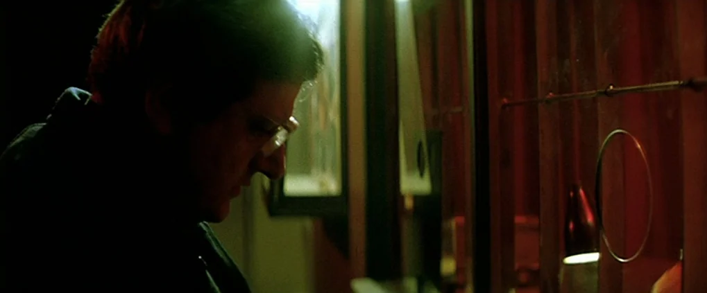 Anguish (1987) movie still: medium — A dark ticket booth interior contains a glass pane with a circular opening. A man in glasses stands in profile…
