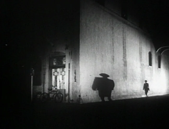 Dementia (Daughter of Horror) (1955) movie still: wide — A dark urban corner features a tall, smooth building wall illuminated by harsh lighting. A…