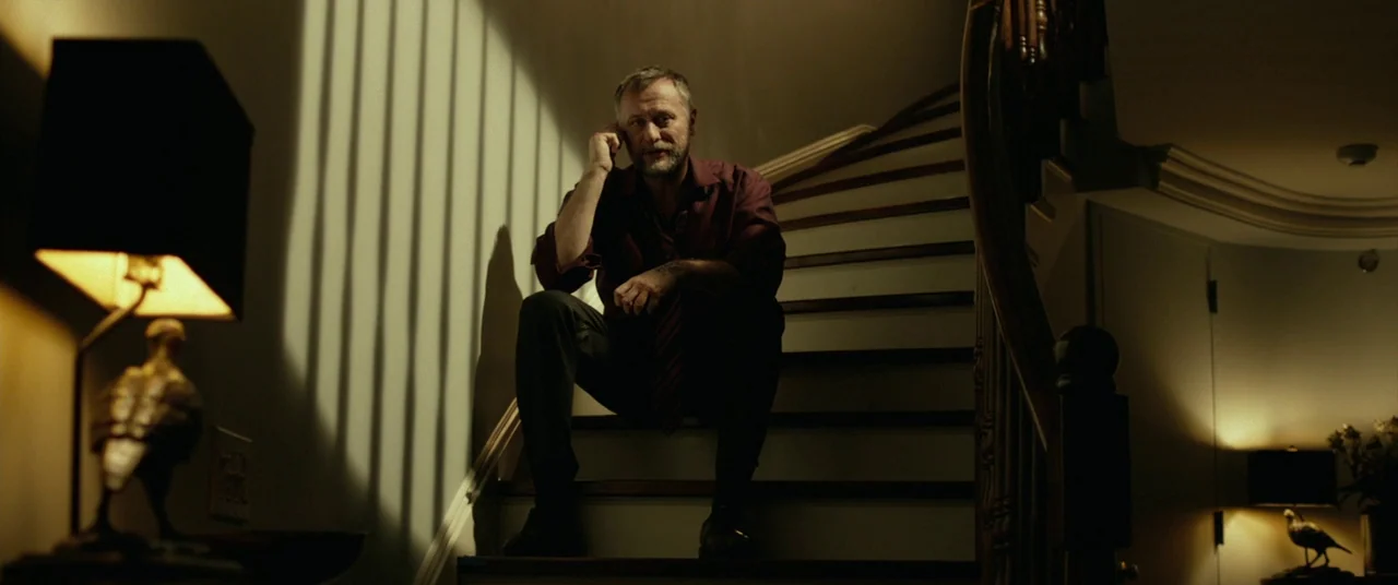 John Wick (2014) movie still: fullbody — A man sits on a wooden staircase in a dimly lit residence at night. He is speaking on a mobile phone while…