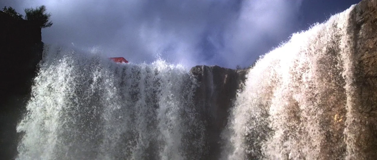 Moonraker (1979) movie still: wide — A wide waterfall drops over a steep, weathered rocky cliff face into empty space. A red object sits near the…