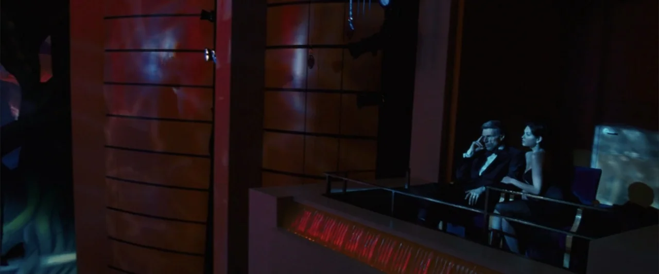 Catwoman (2004) movie still: wide — A dark, modern nightclub balcony with red neon light streaks reflecting on wall panels. A man and a woman in…