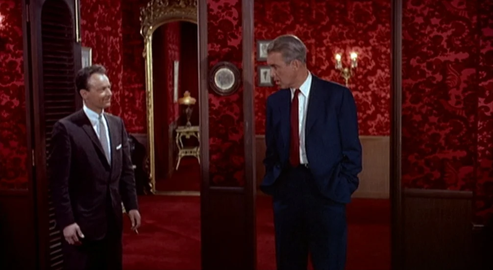 Vertigo (1958) movie still: fullbody — A lavish room features vibrant red patterned wallpaper and matching plush carpeting throughout. Two men in…