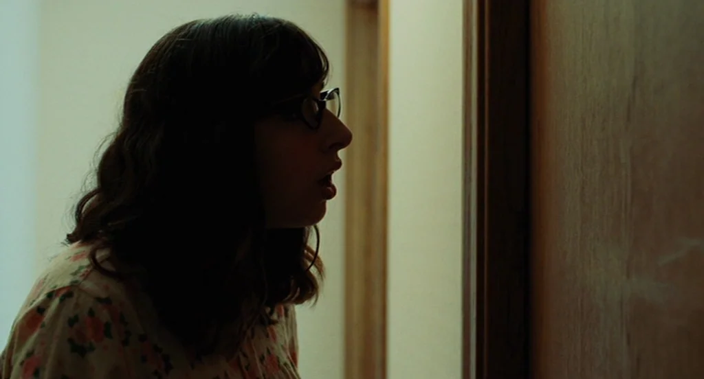 A Serious Man (2009) movie still: closeup — A woman stands in a dim indoor area near a wooden door frame. She looks ahead with her mouth slightly…