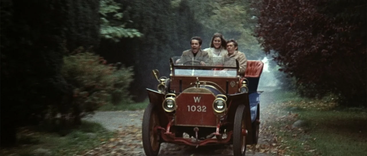 Duck, You Sucker (1971) movie still: medium — A rural road lined with dense dark trees and fallen leaves in autumn. Three people sit inside a red…