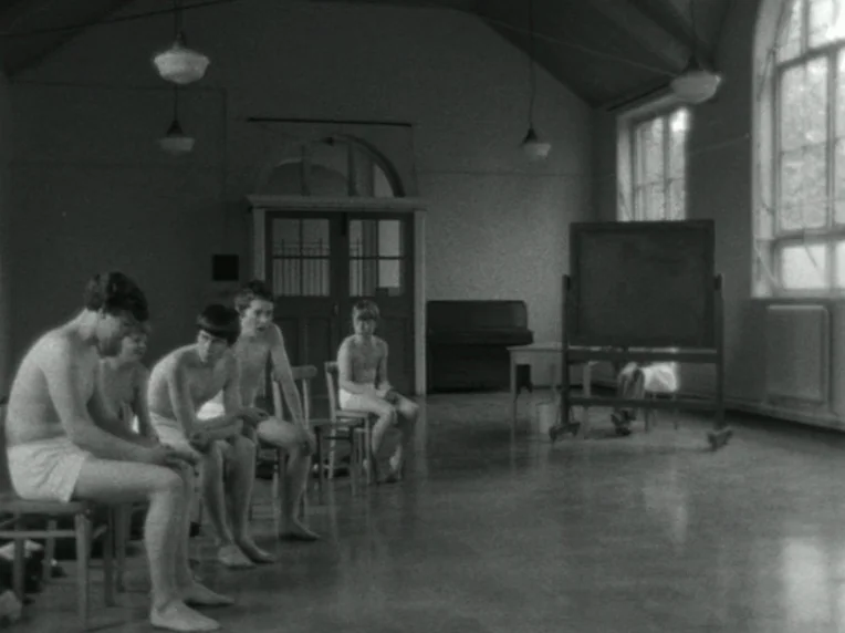 Children (1976) movie still: medium — A high-ceilinged room with large windows, simple wooden chairs, and a freestanding blackboard. Four boys sit…