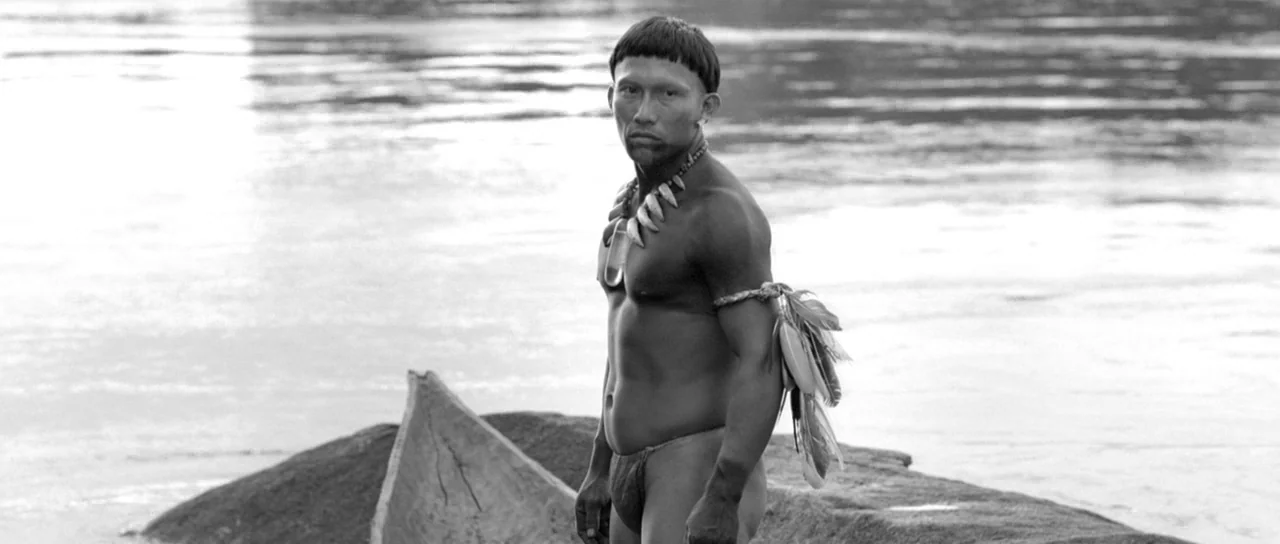 Embrace Of The Serpent (2015) movie still: medium — A riverbank features a small wooden canoe resting on a flat stone surface. A man wearing…