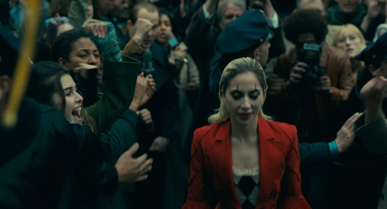 Joker: Folie à Deux (2024) movie still: medium — A dense crowd of people presses in from all sides on a city street. A woman in a red blazer walks…
