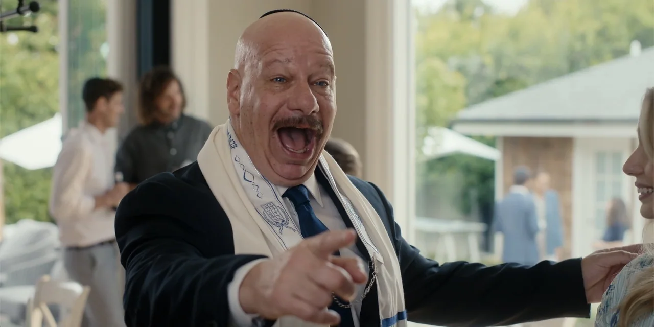 Ricky Stanicky (2024) movie still: medium — An interior reception space features large windows overlooking an exterior patio area. An older bald man…