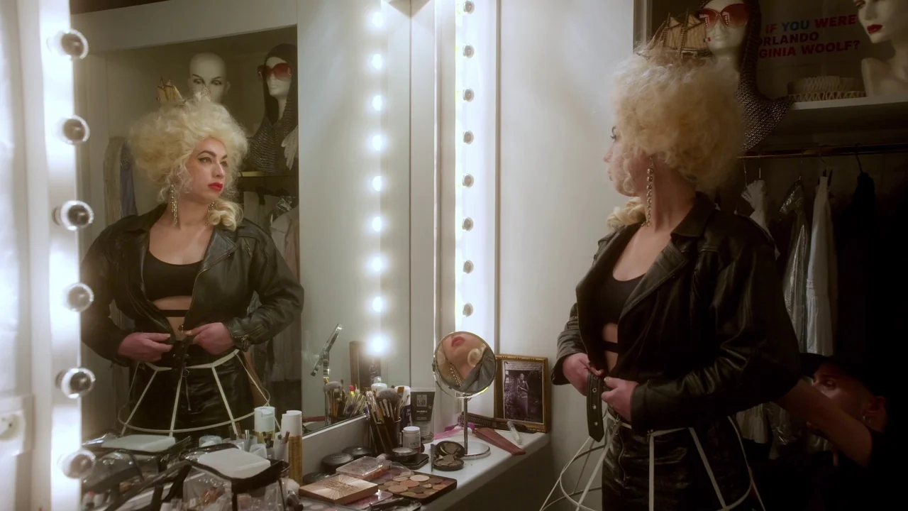Orlando: My Political Biography (2023) movie still: medium — A well-lit dressing area contains a vanity mirror bordered by bright, vertical bulbs.…