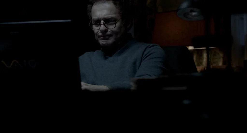 Mama (2013) movie still: medium — A dark, dimly lit home workspace with a man sitting before a computer. The man stares directly at the glowing…