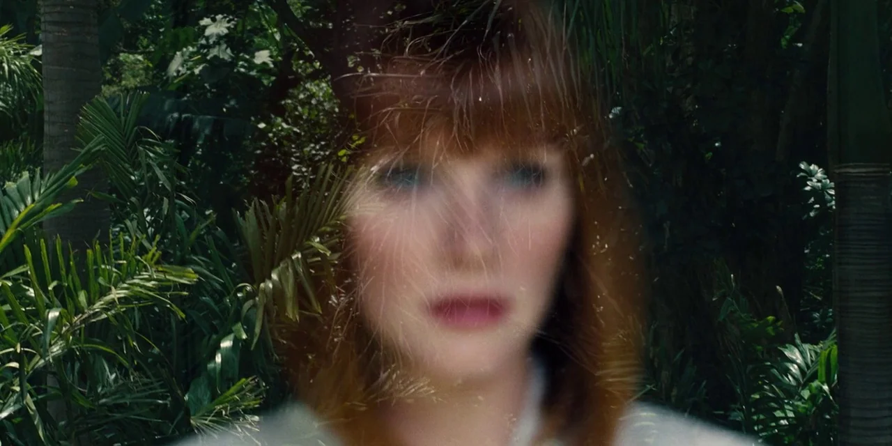 Jurassic World (2015) movie still: closeup — A woman stands behind dense tropical foliage that creates a blurred overlay. She looks towards the…