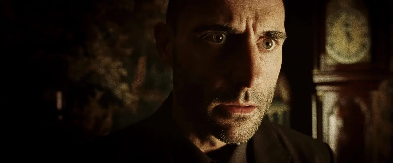 Anna (2013) movie still: closeup — A dimly lit room contains a grandfather clock in the blurred background. A middle-aged man stares directly…