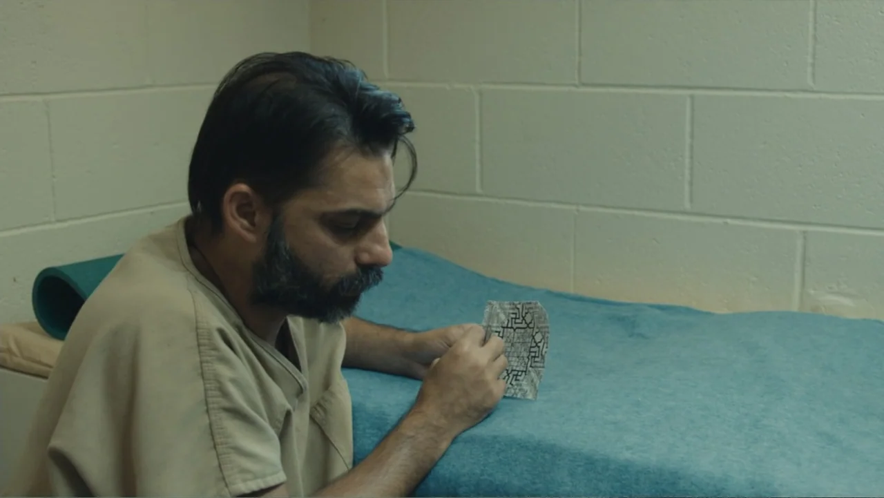 Camp X-Ray (2014) movie still: medium — A stark prison cell features beige painted cinderblock walls and a bed. A bearded man in a tan scrub top…