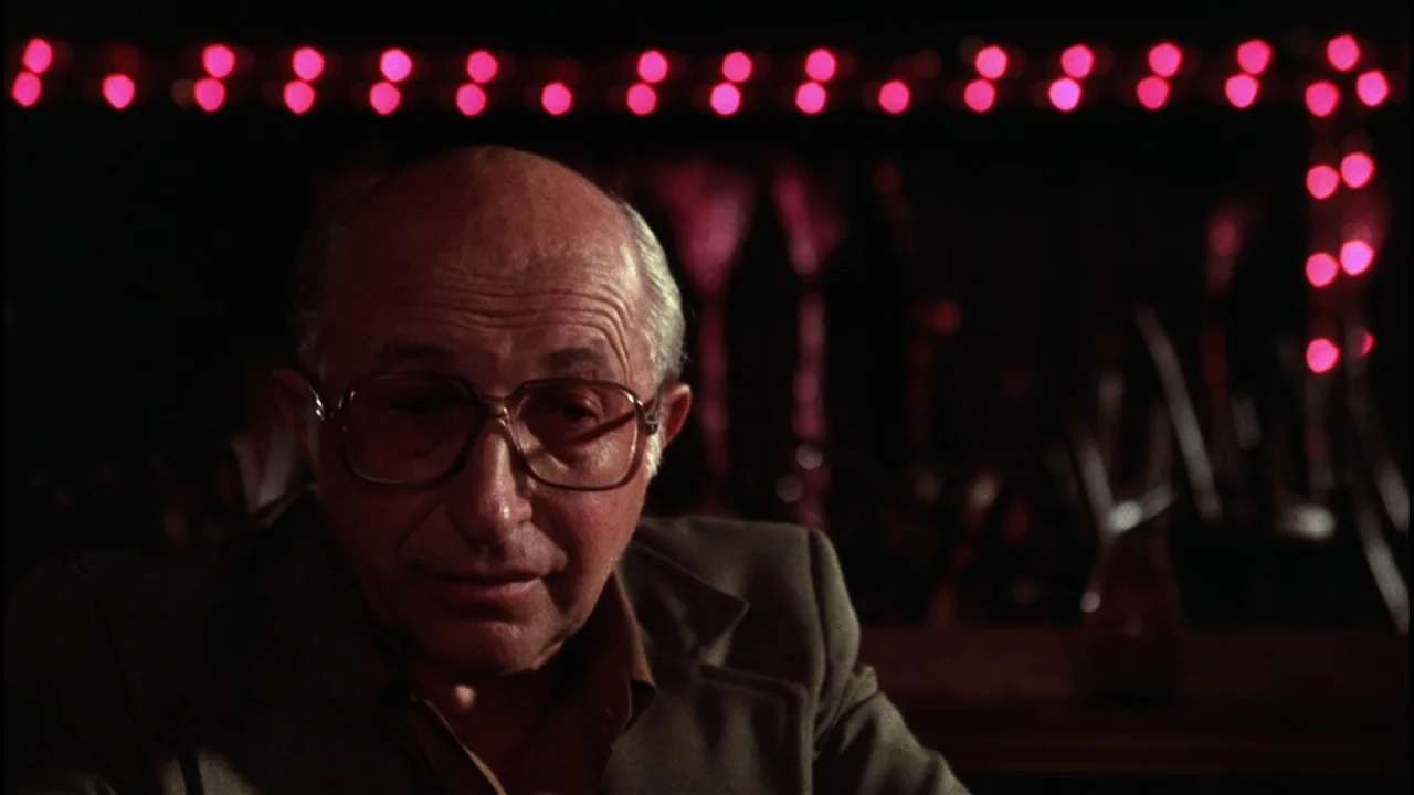 Star 80 (1983) movie still: medium — A dimly lit bar interior features a wall of glowing red neon lights. An older man with glasses sits in the…