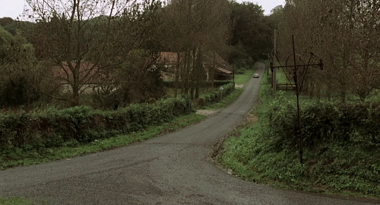 The Diving Bell and the Butterfly (2007) movie still: establishing — A rural paved road winds through a lush, green countryside with dense trees. A…