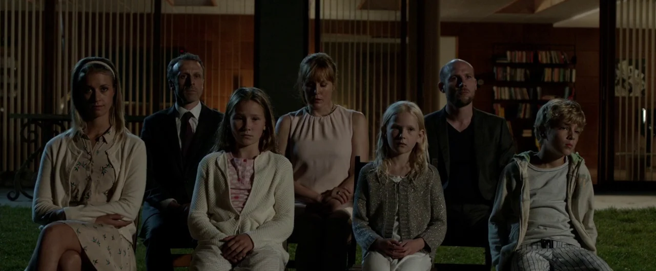 Borgman (2013) movie still: medium — A group of six people sits in a line on a darkened patio at night. Behind them is a large glass-walled…