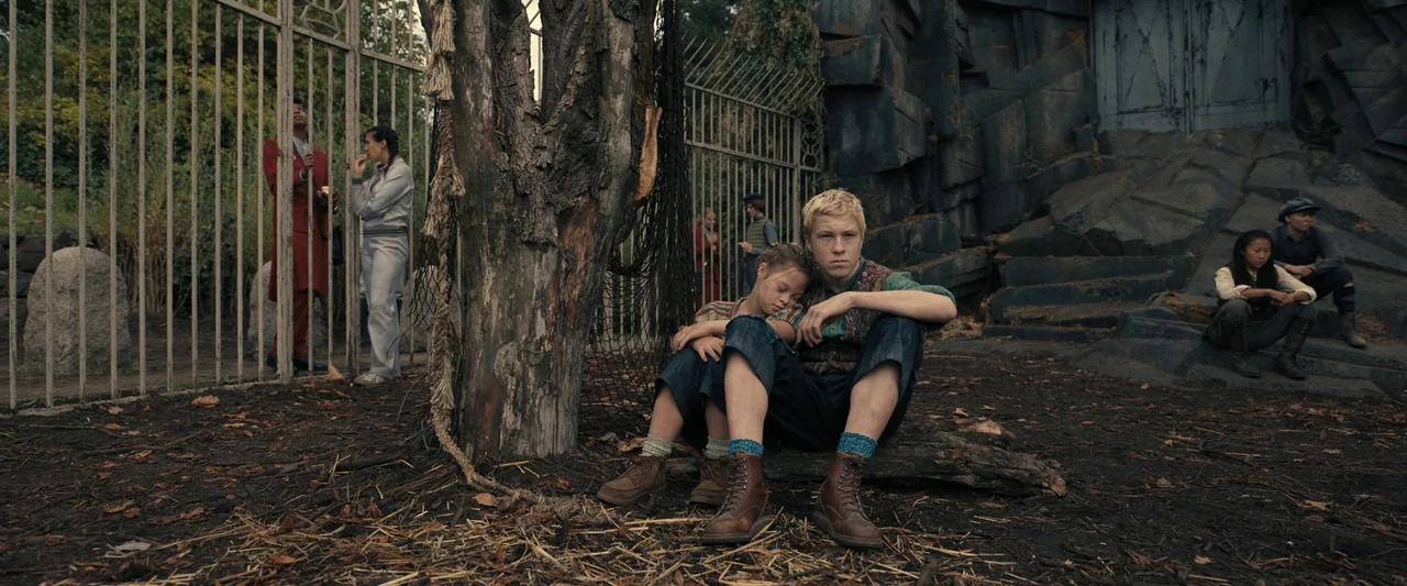 The Hunger Games: The Ballad of Songbirds & Snakes (2023) movie still: wide — A group of children occupy a dirt-covered area near a tall metal…
