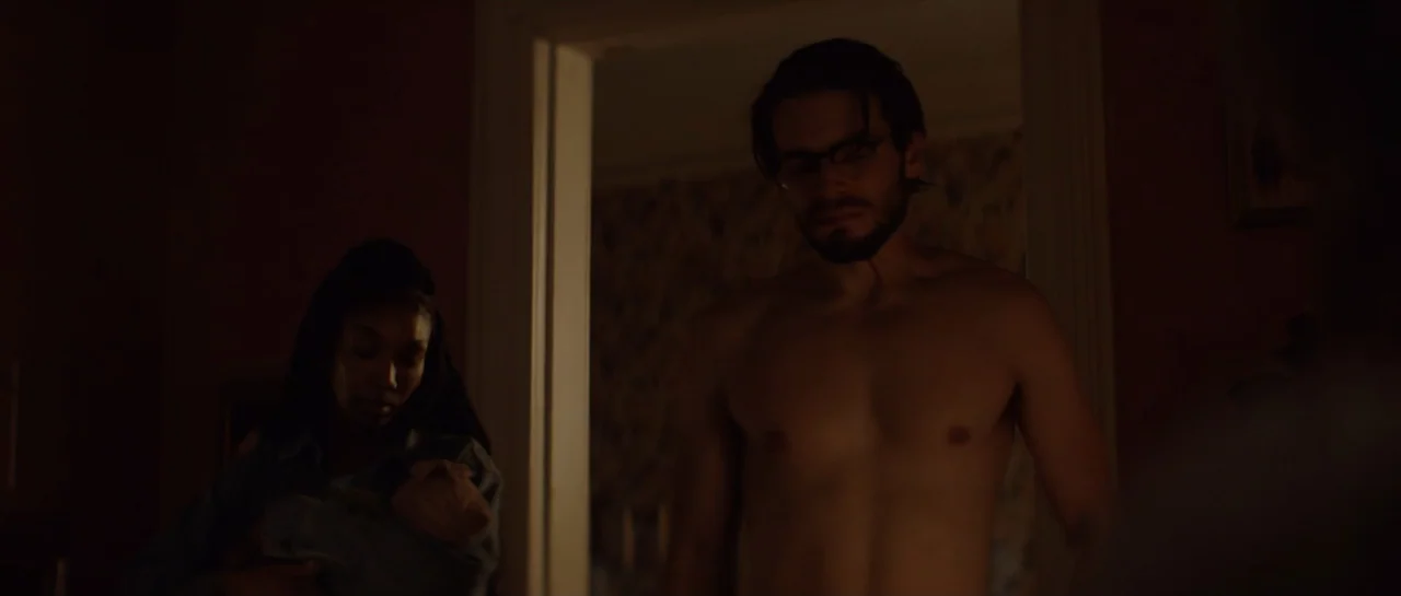 The Front Room (2024) movie still: medium — A dimly lit room with patterned wallpaper visible through an open wooden doorway. A shirtless man moves…