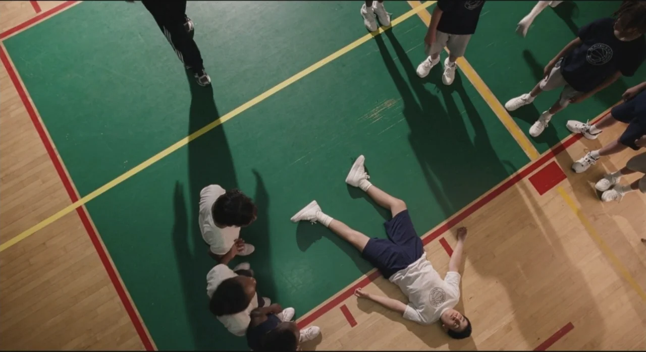 St. Vincent (2014) movie still: wide — A high-angle view of a gymnasium floor with green and yellow painted markings. A group of teenagers in…