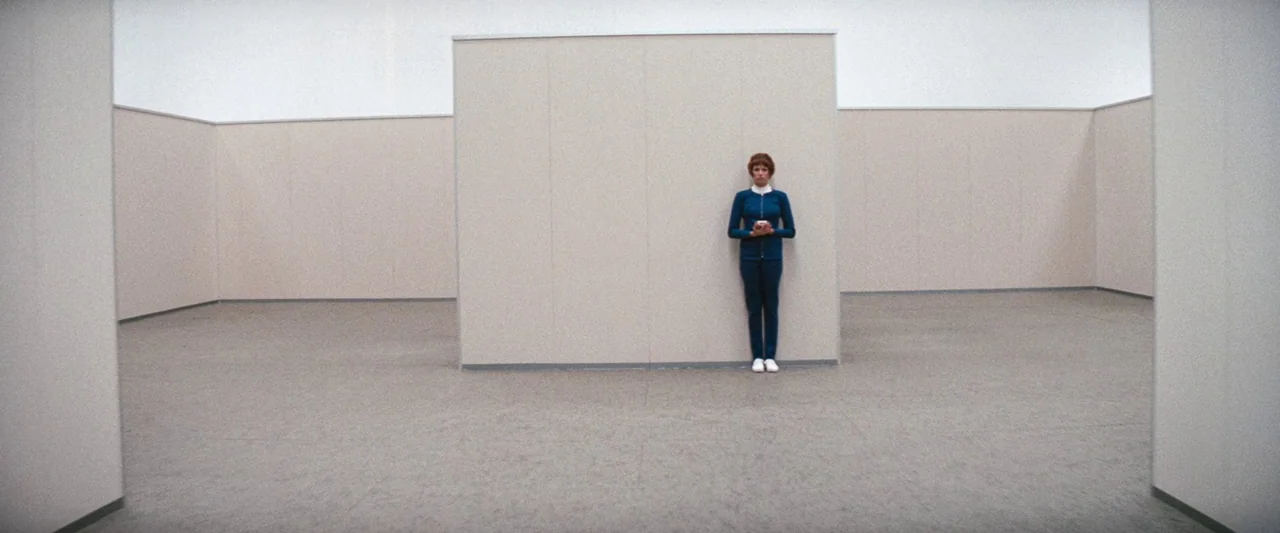 Westworld (1973) movie still: wide — A minimalist gallery room features light beige walls and grey carpeted flooring. A woman in a blue uniform…