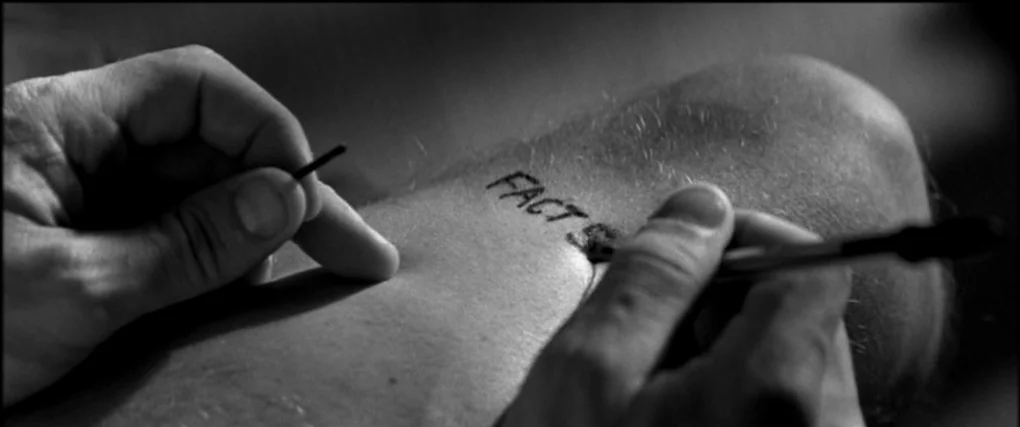 Memento (2000) movie still: closeup — A close view of a person's thigh with the word FACT written on it. A hand holds a ballpoint pen and is in the…