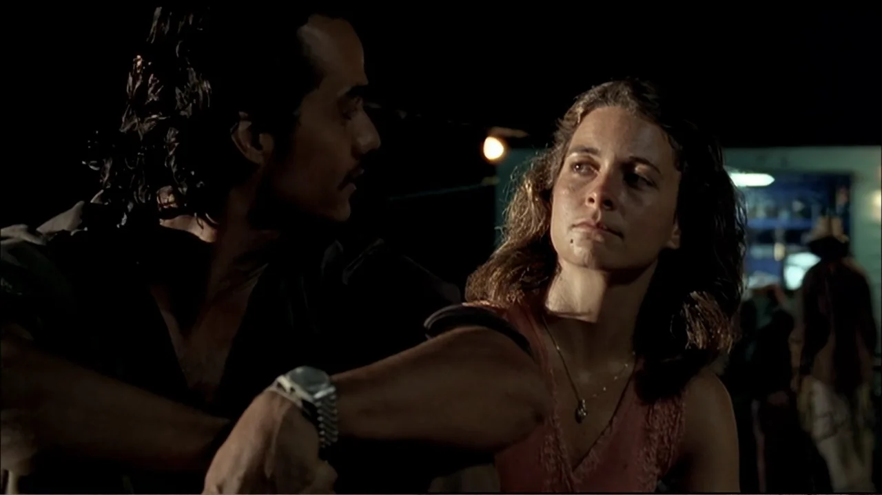 In the Middle of the World (1990) movie still: medium — An outdoor space at night with dark surroundings and a illuminated background area. A man…
