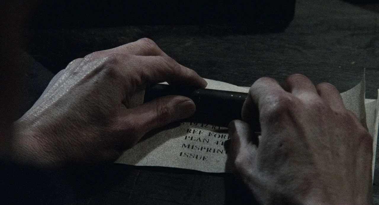 1984 (1984) movie still: closeup — Two hands rest on a dimly lit wooden surface while holding a document. The paper contains printed text…