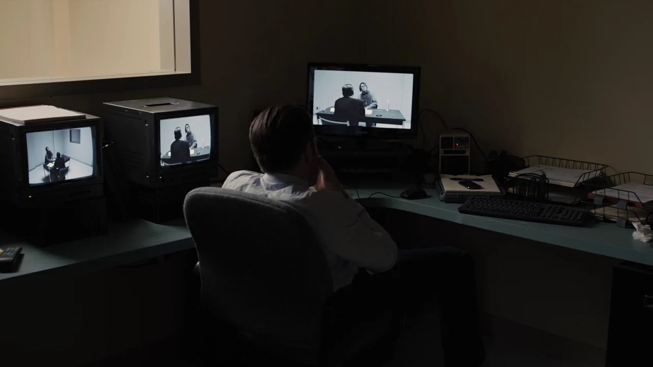 Prisoners (2013) movie still: medium — A dimly lit surveillance room contains multiple monitors displaying images of a separate room. A lone man…