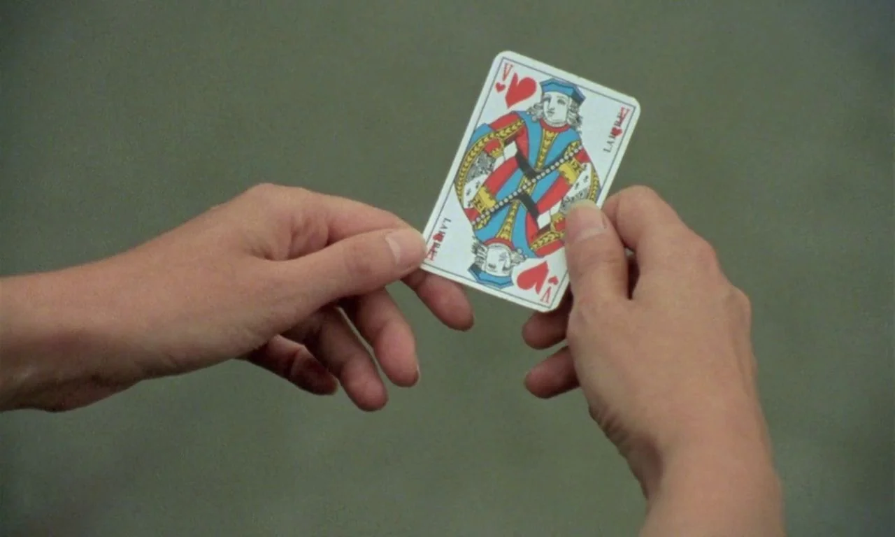 The Green Ray (1986) movie still: closeup — A playing card is held between two hands against a plain olive-grey backdrop. The card displays a…