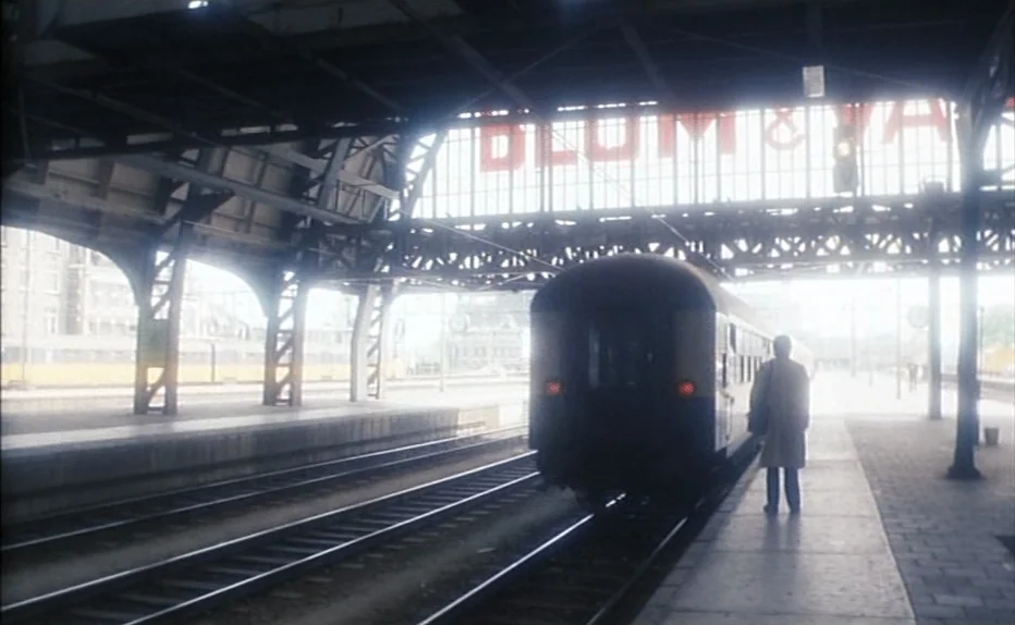The Fourth Man (1983) movie still: wide — A covered train platform with large iron support beams and high glass ceilings. A man stands alone on the…